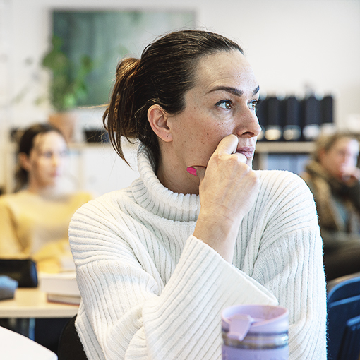Socialpedagog_510_web kopiera Kvinna med mörkt hår och ljus tröja sitter i en lektionssal med fler personer.