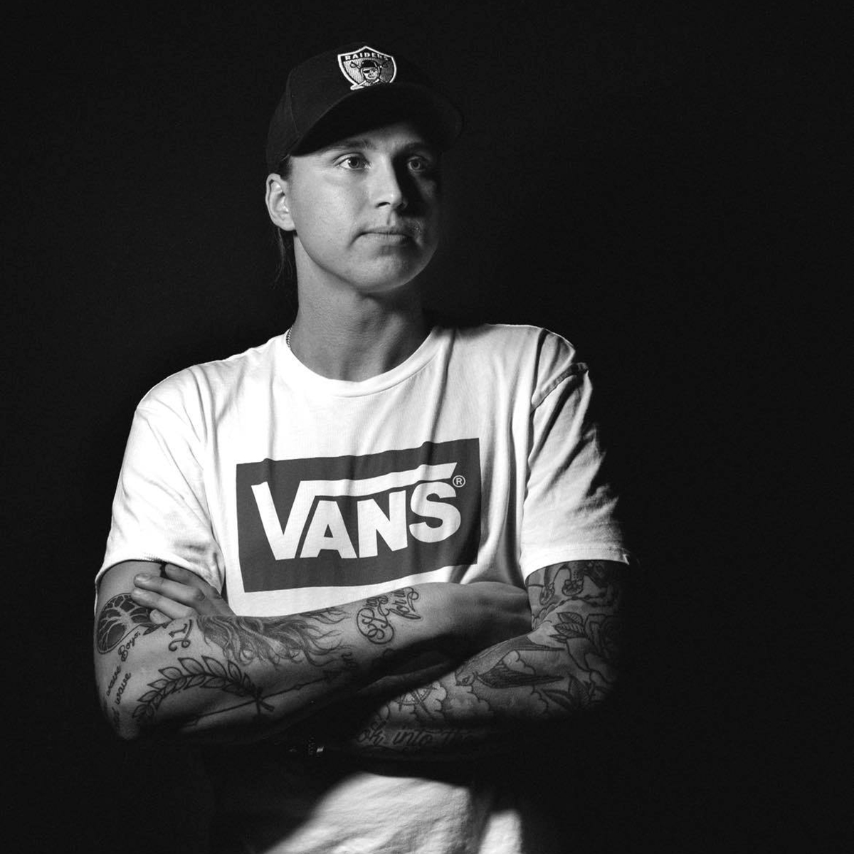 Svartvit bild på en ung man med keps och tatueringar på armarna.